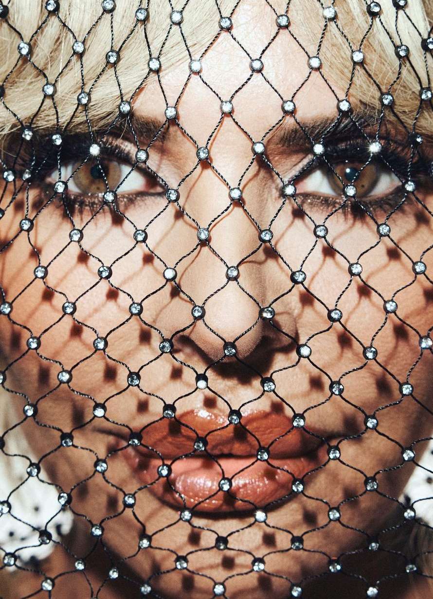 Close-up portrait of a woman's face partially obscured by a black net veil adorned with rhinestones, focusing on her eyes and lips.