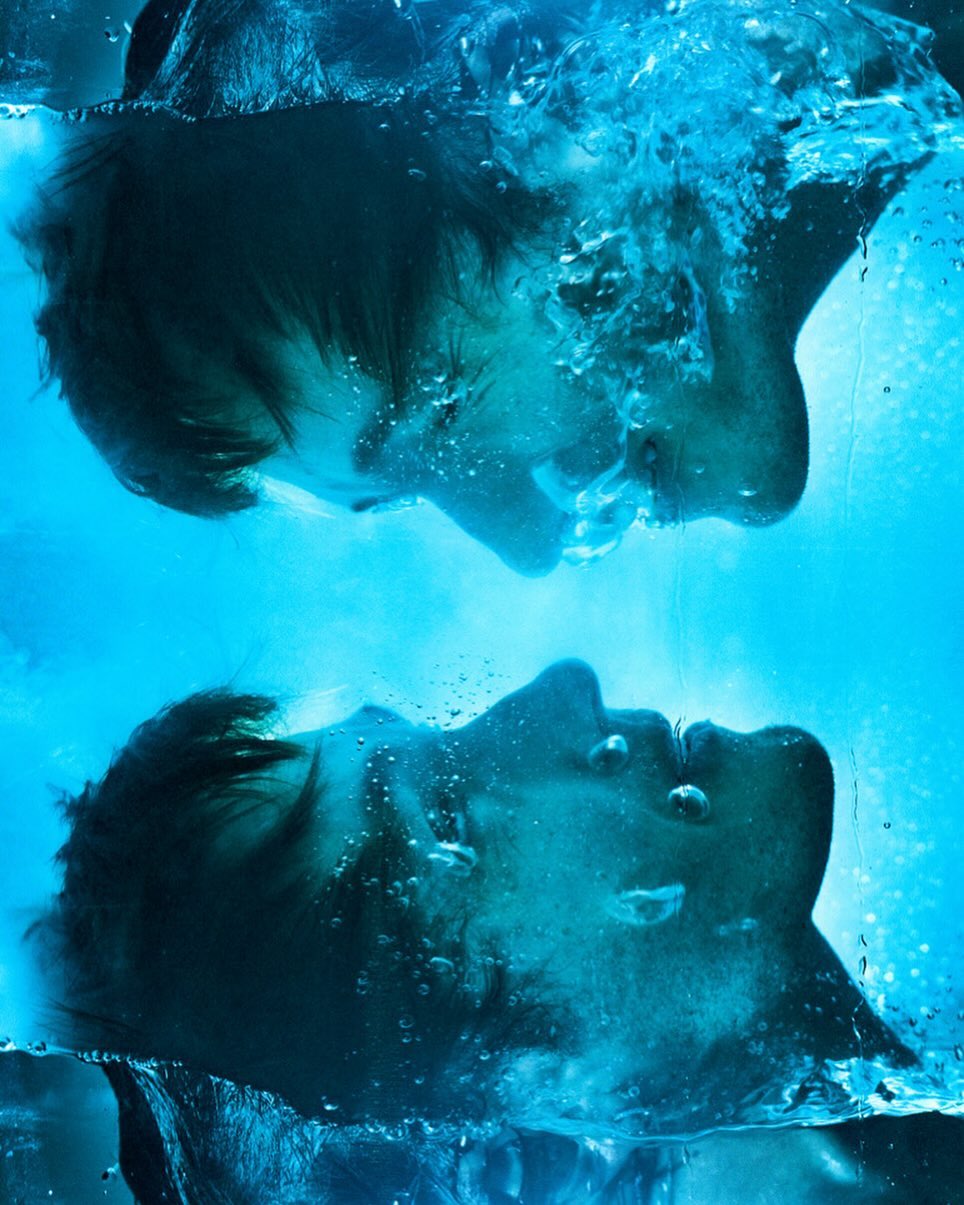 Underwater portrait of two adult males facing each other, with bubbles creating abstract shapes in bright teal-colored water.