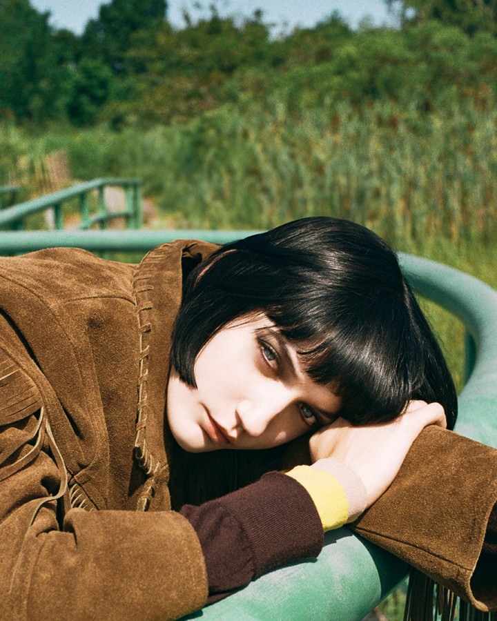 Close-up shot of a young woman with dark hair lying on a green metal structure in a field with trees in the background, wearing a brown fringe jacket.
