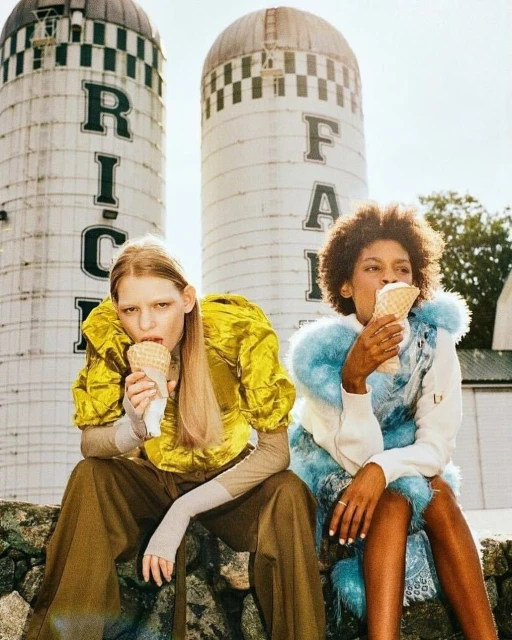 Two stylish women enjoy ice cream cones while seated on a stone wall with silos and farm buildings in the background under the sun.