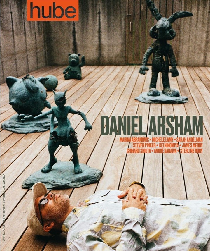 Man lies near art sculptures on a wooden surface for magazine cover; sculpture of cartoon characters and geometric gray wall backdrop.