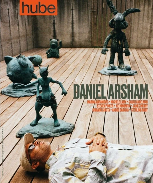 Man lies near art sculptures on a wooden surface for magazine cover; sculpture of cartoon characters and geometric gray wall backdrop.