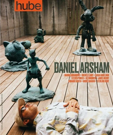 Man lies near art sculptures on a wooden surface for magazine cover; sculpture of cartoon characters and geometric gray wall backdrop.