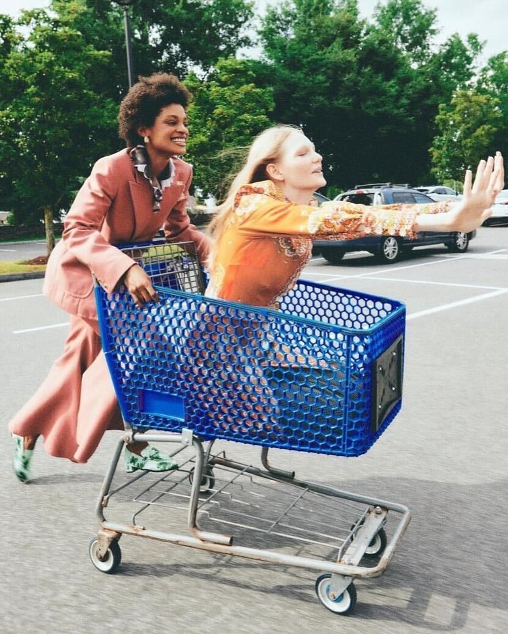 Two women in a parking lot, one pushing a blue shopping cart with the other riding inside, both smiling, showcasing fun and street style.