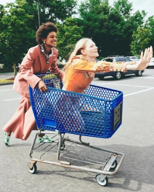 Two women in a parking lot, one pushing a blue shopping cart with the other riding inside, both smiling, showcasing fun and street style.
