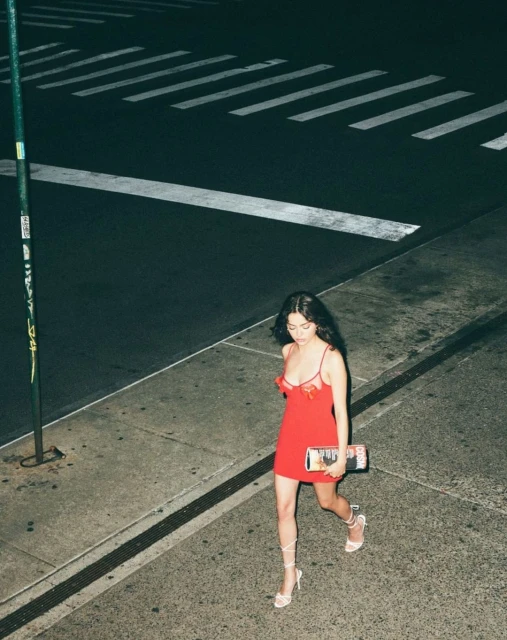 A woman in a red dress and heels strides across a crosswalk at night, holding a magazine or booklet in her hand in an urban setting.