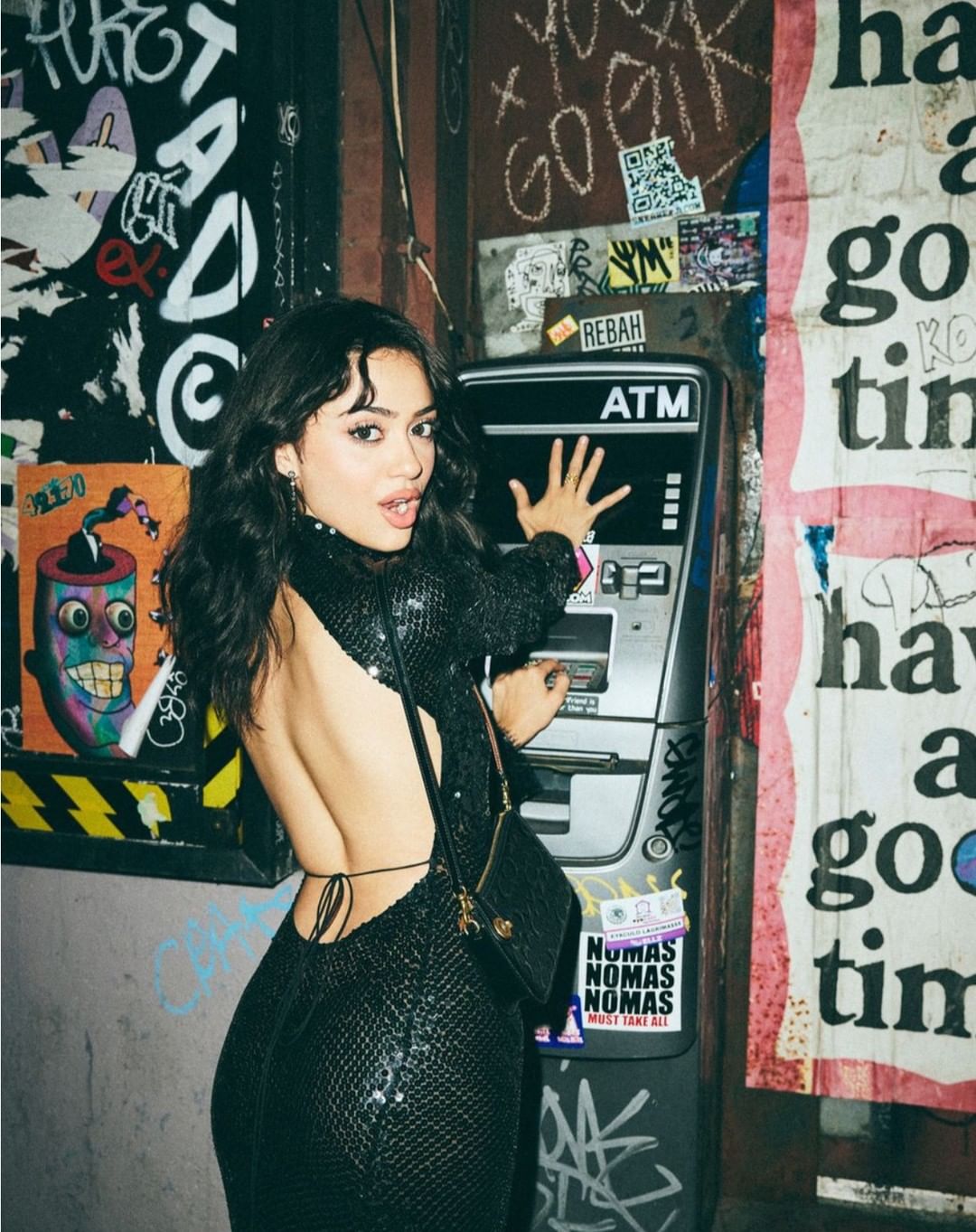 A woman in a black dress uses an ATM on a city street, surrounded by graffiti and posters, in a candid and vibrant urban scene.