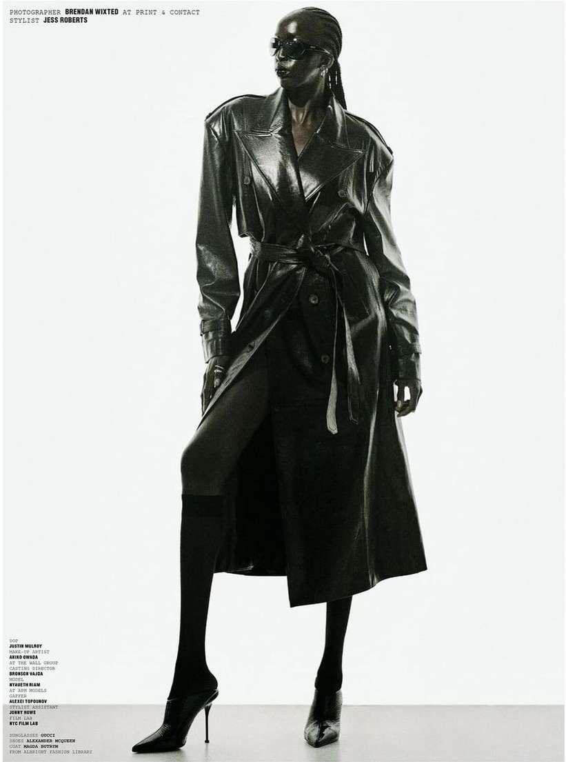 Full-length studio shot of a Black woman wearing a black shiny trench coat, black high-heeled shoes and sunglasses against a white background.