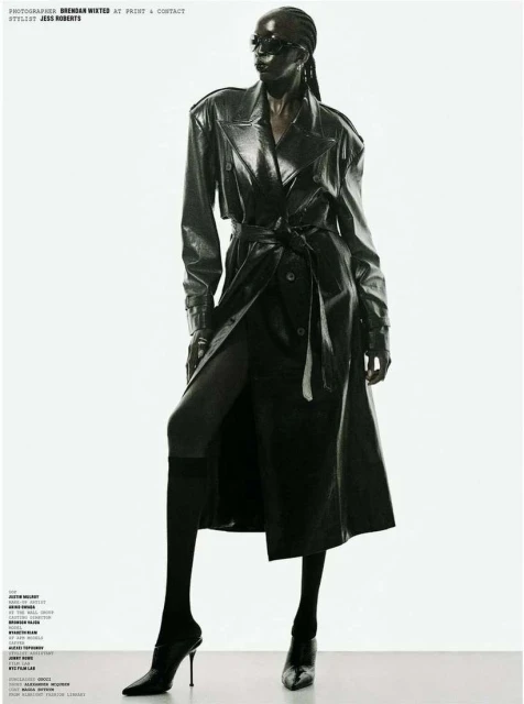 Full-length studio shot of a Black woman wearing a black shiny trench coat, black high-heeled shoes and sunglasses against a white background.