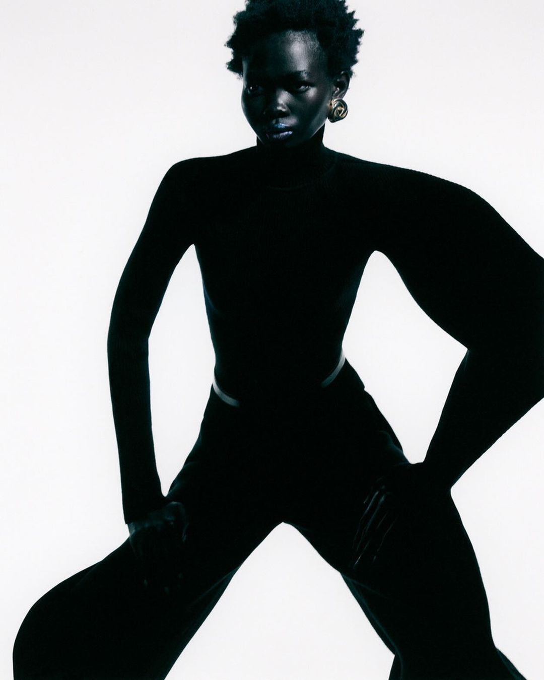 A full-length studio portrait of an adult Black woman with short natural hair, wearing all-black clothing against a bright white backdrop.