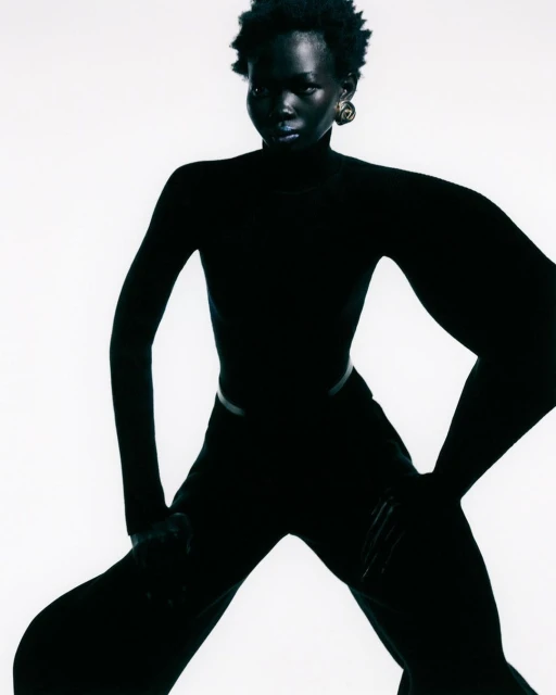 A full-length studio portrait of an adult Black woman with short natural hair, wearing all-black clothing against a bright white backdrop.