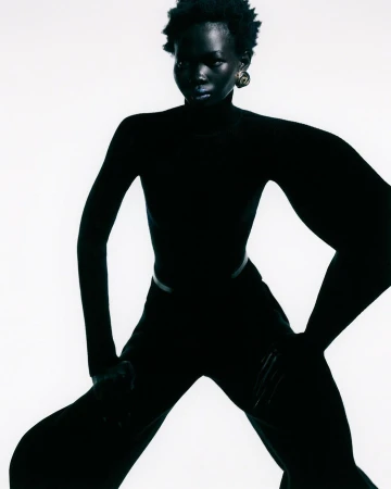 A full-length studio portrait of an adult Black woman with short natural hair, wearing all-black clothing against a bright white backdrop.