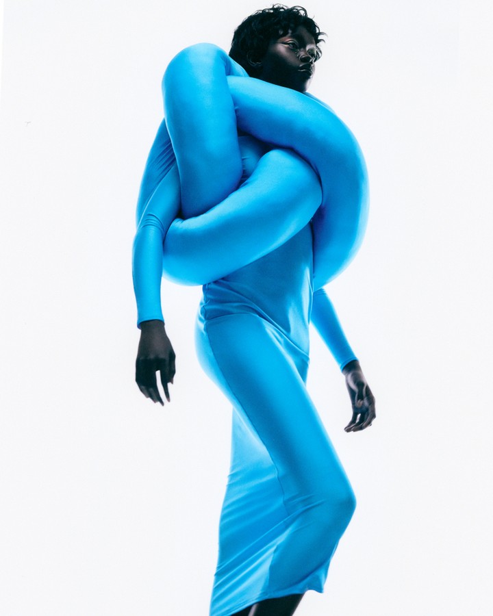 Woman in long-sleeve turquoise dress and inflated tube around shoulders poses on white backdrop in studio lighting for fashion editorial.