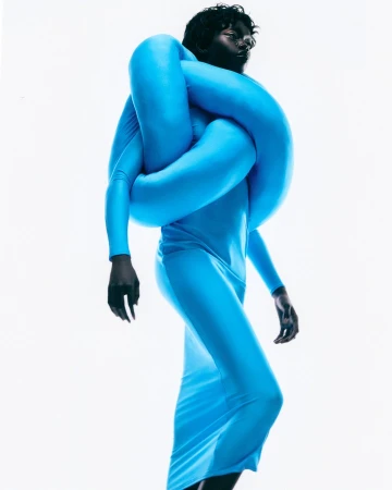 Woman in long-sleeve turquoise dress and inflated tube around shoulders poses on white backdrop in studio lighting for fashion editorial.