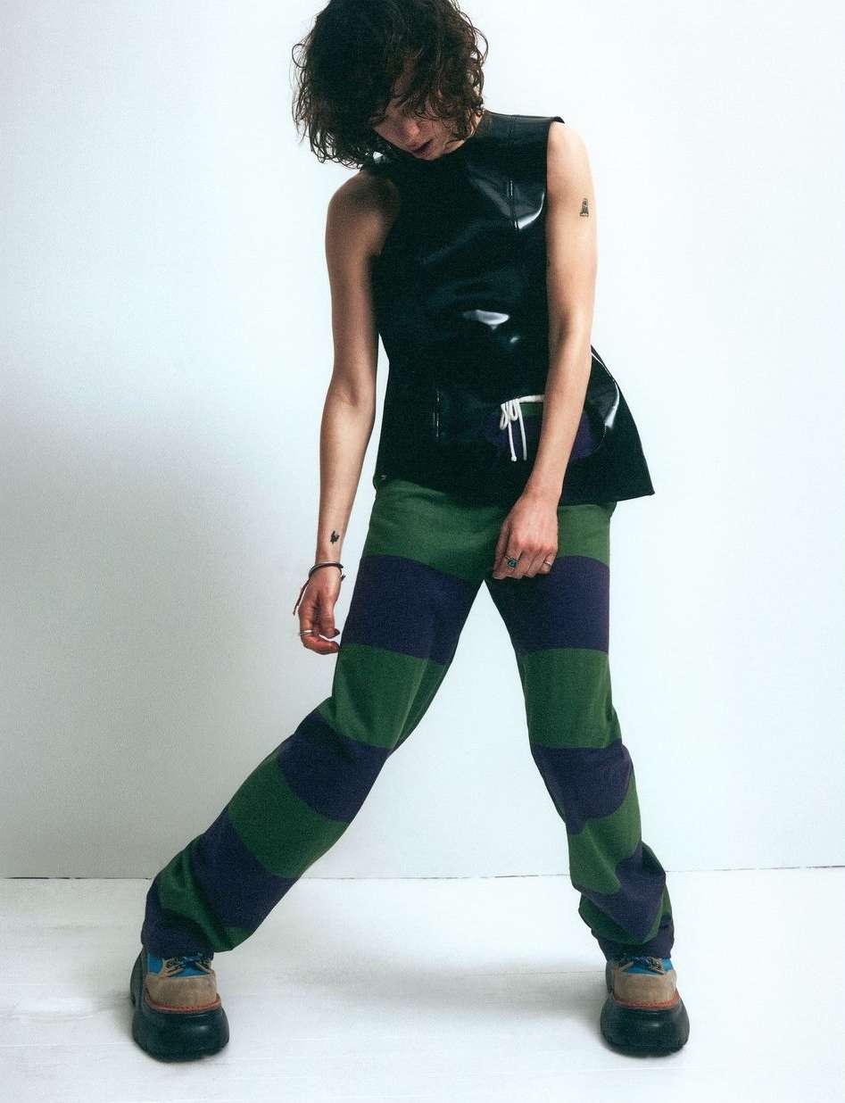Non-binary person in sleeveless black top and striped pants against a white background, posing in a fashion forward stance.