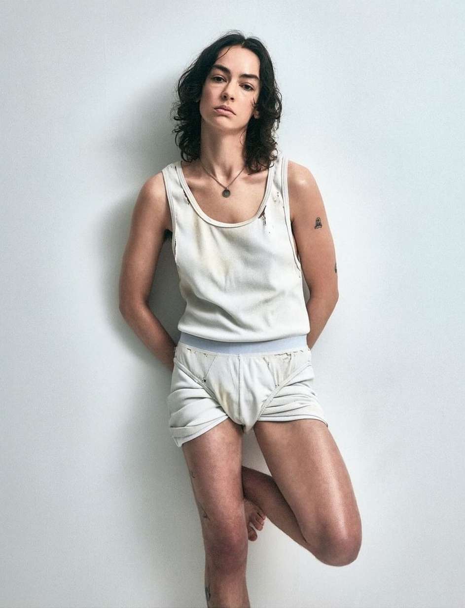 Woman with dark hair wearing a white tank top and shorts leaning against a plain light wall