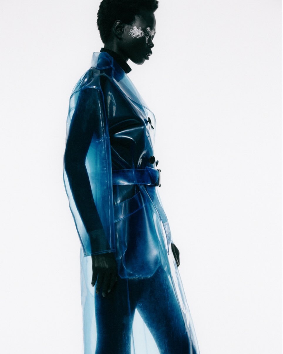 Fashionable Black woman in a translucent blue trench coat with silver eye makeup against a stark white backdrop in a studio setting.