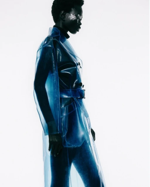 Fashionable Black woman in a translucent blue trench coat with silver eye makeup against a stark white backdrop in a studio setting.