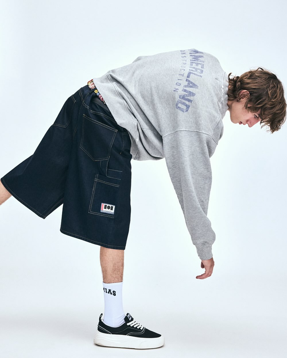 Young adult wearing grey Timberland sweatshirt, black denim shorts, white socks, and black and white platform sneakers against white background.