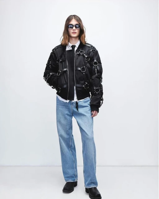 Full-length shot of a stylish adult male wearing a black bomber jacket, light blue jeans, and black boots, set against a plain white backdrop.