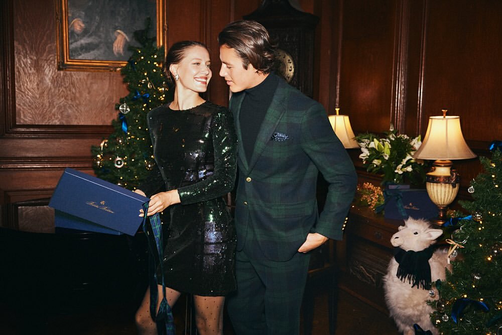 Elegant couple in evening wear standing near Christmas tree indoors. Man in plaid suit, woman in sequin dress holding gift boxes.