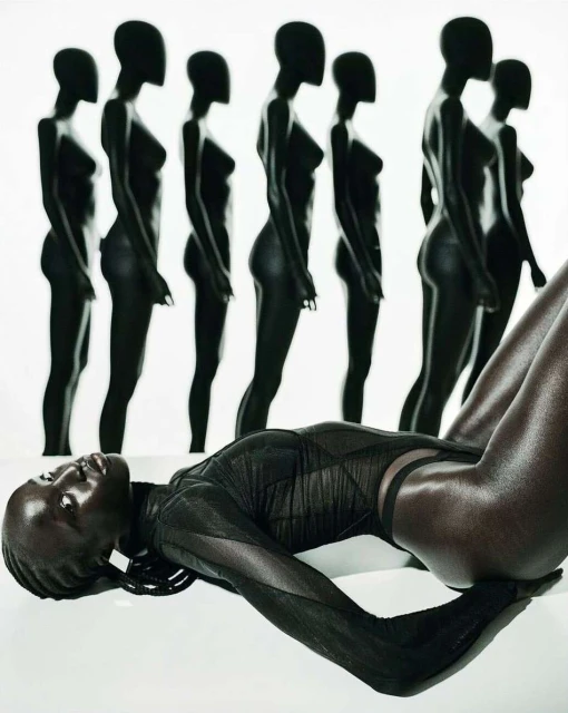 Striking artistic fashion shot of an African woman lying down with mannequins in the background, highlighting form and texture in black tones.