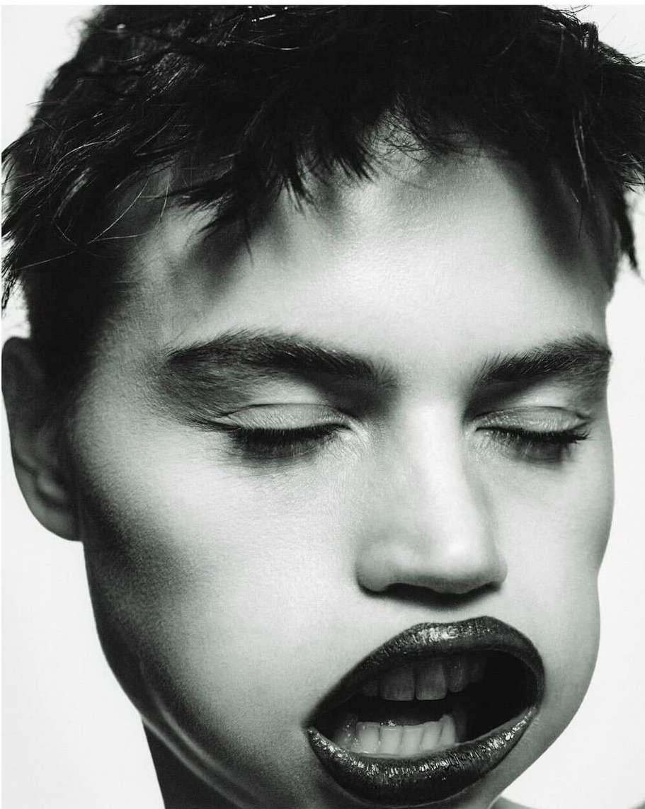 Monochromatic close-up of a young adult woman's face with her eyes closed and mouth open, revealing teeth and dark lipstick, against a white background.