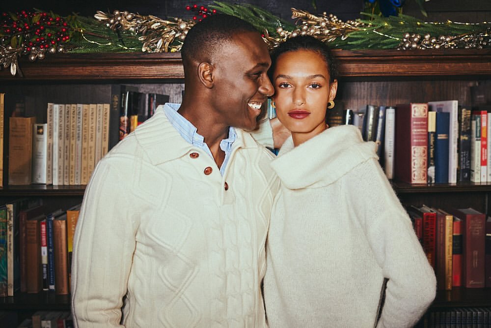Indoor portrait of a smiling Black man and mixed-race woman wearing cream-colored sweaters, posing in front of a bookshelf.