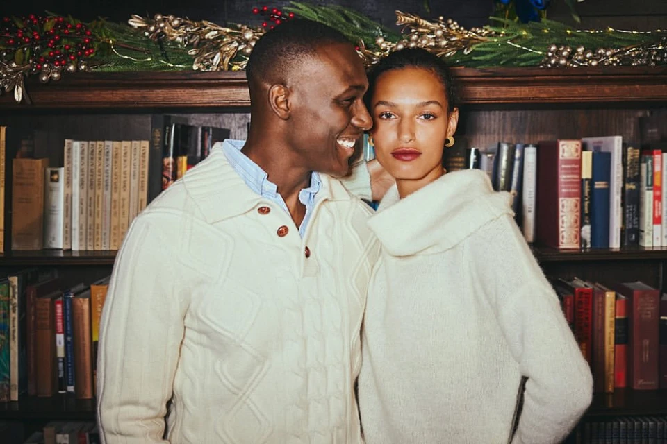 Indoor portrait of a smiling Black man and mixed-race woman wearing cream-colored sweaters, posing in front of a bookshelf.