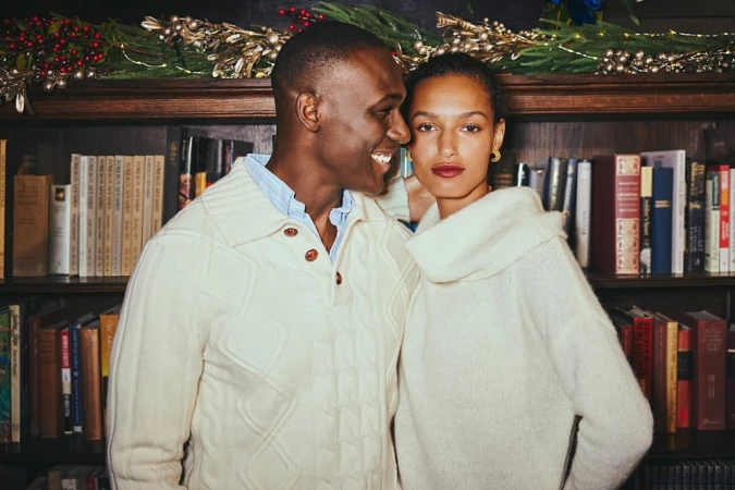 Indoor portrait of a smiling Black man and mixed-race woman wearing cream-colored sweaters, posing in front of a bookshelf.