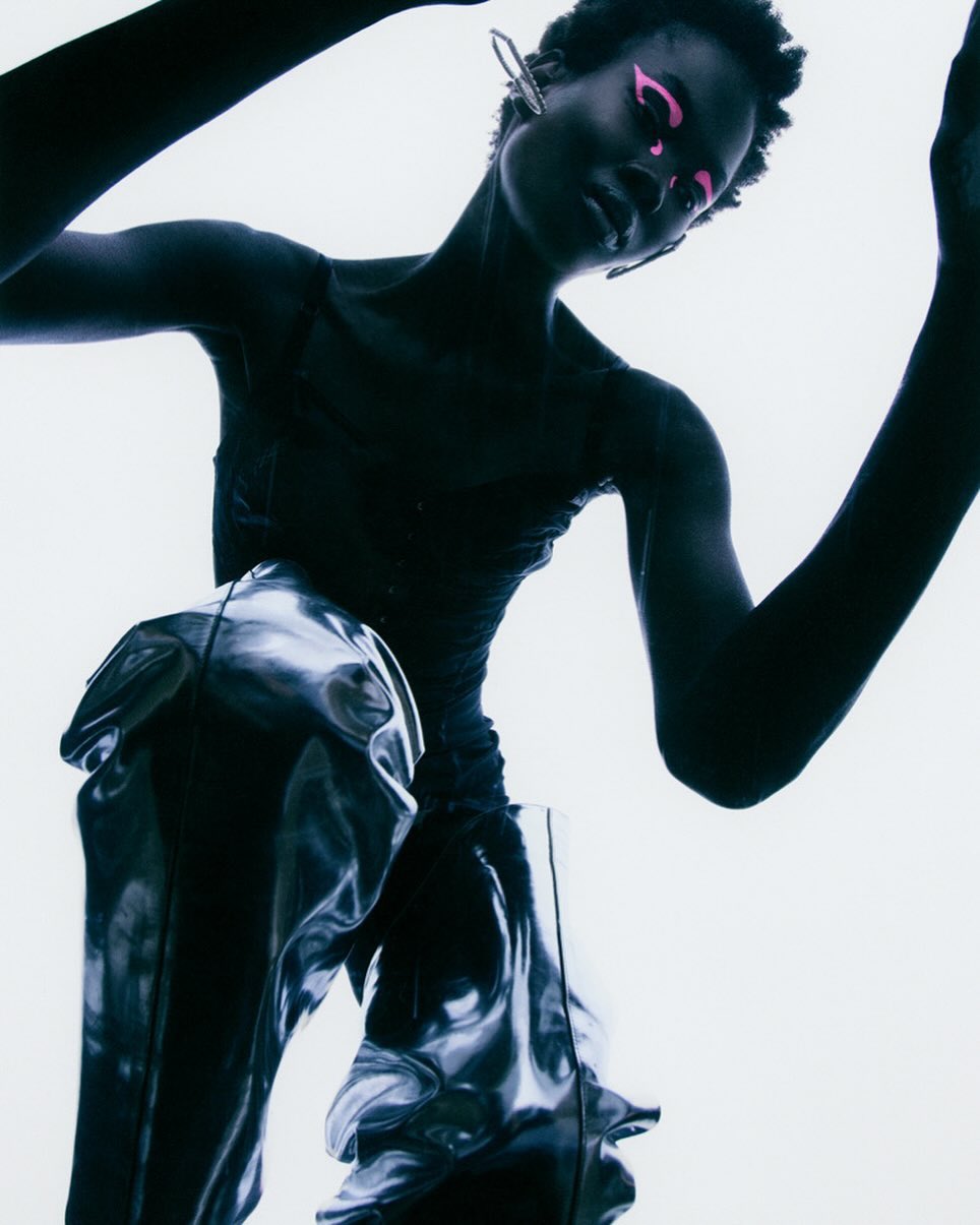 Fashion image of a Black female model with pink makeup wearing a metallic-looking skirt against a bright, high-key background.
