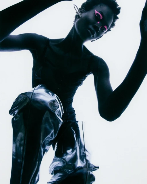 Fashion image of a Black female model with pink makeup wearing a metallic-looking skirt against a bright, high-key background.
