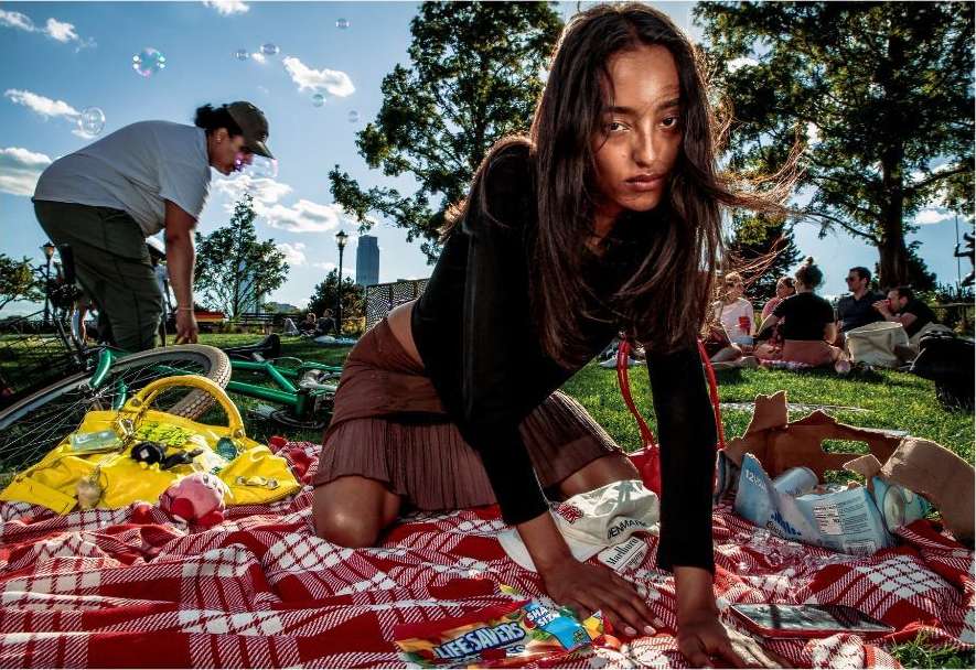 Woman kneeling on a red and white blanket, picnic style, in a park with other people, green grass, and bubbles in the air.