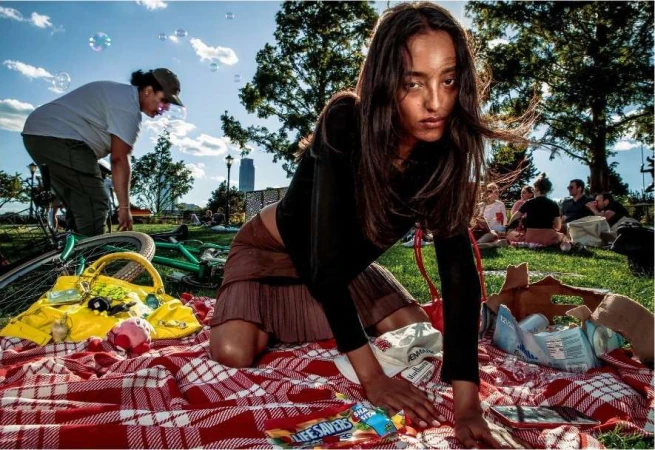 Woman kneeling on a red and white blanket, picnic style, in a park with other people, green grass, and bubbles in the air.
