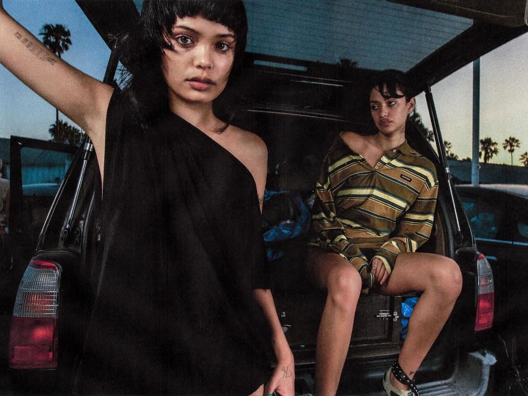 Two young women pose in the open trunk of a black car at twilight. One woman wears a draped black dress; the other sits in a striped top.