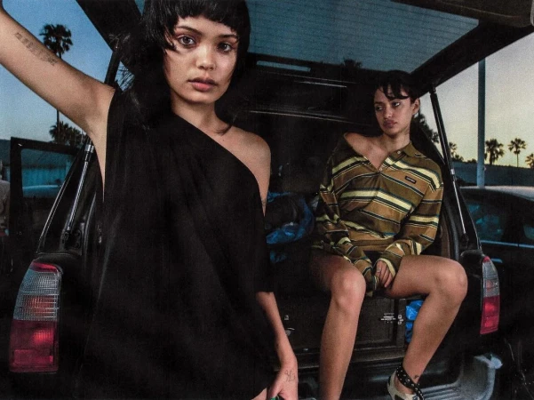Two young women pose in the open trunk of a black car at twilight. One woman wears a draped black dress; the other sits in a striped top.