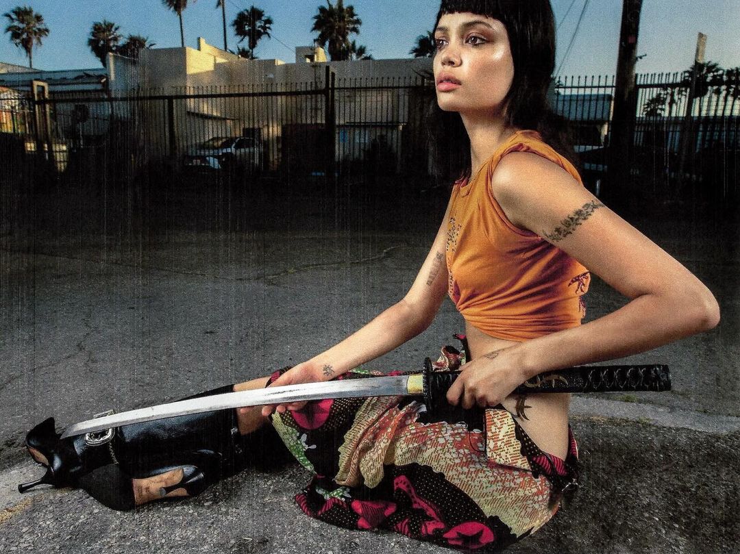 Woman poses on the street with a katana, wearing a cropped orange top and patterned skirt, with palm trees and urban buildings behind her.