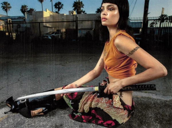 Woman poses on the street with a katana, wearing a cropped orange top and patterned skirt, with palm trees and urban buildings behind her.