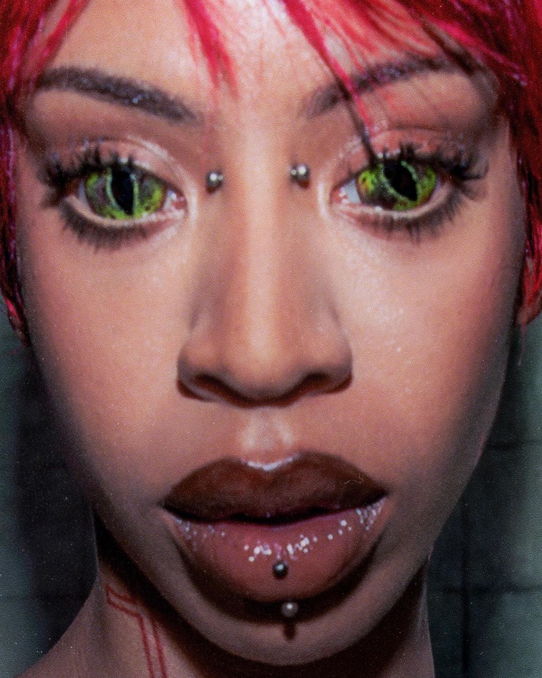 Close-up portrait of a woman with striking green reptilian eyes, glossy brown lips, and red hair against a dark, textured background.