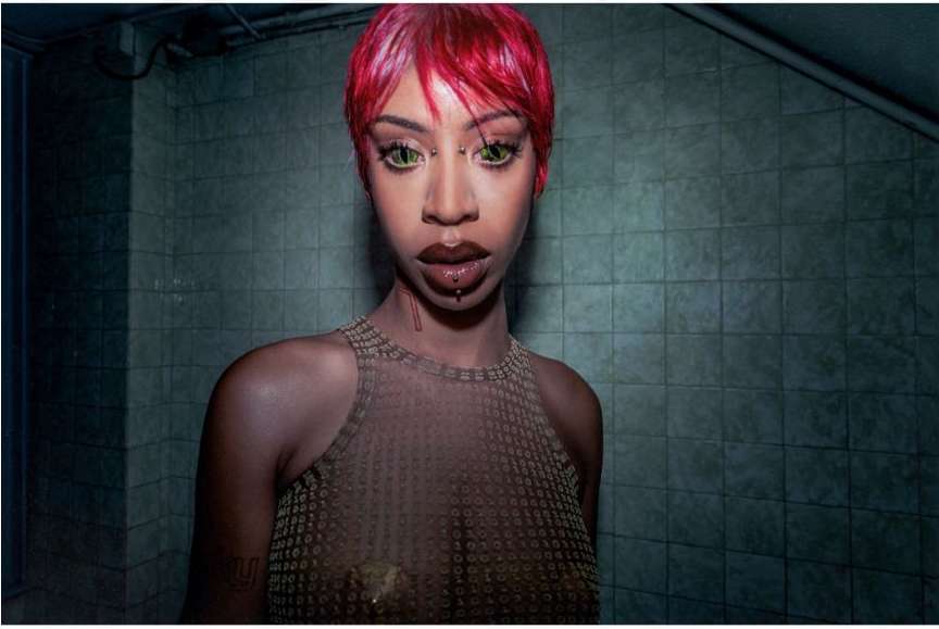 Portrait of a woman with short red hair, light green eyes, and multiple facial piercings, wearing a mesh top in a tiled room.