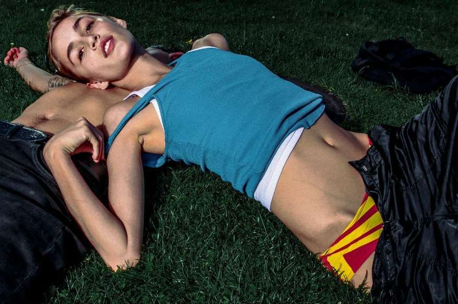 A tan-skinned woman and man lying on green grass, with the woman wearing a blue tank top and yellow bottoms, and the man shirtless.