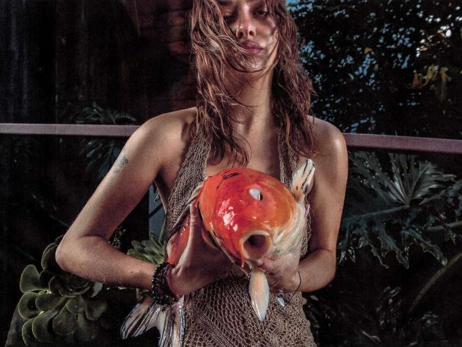 Adult woman holds a large orange and white Koi fish in an outdoor setting with lush foliage in the background, posing for a portrait.