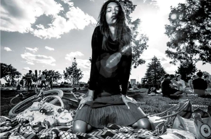 Black and white portrait of a young woman kneeling in a field with bicycles, clouds, and trees visible in the background.