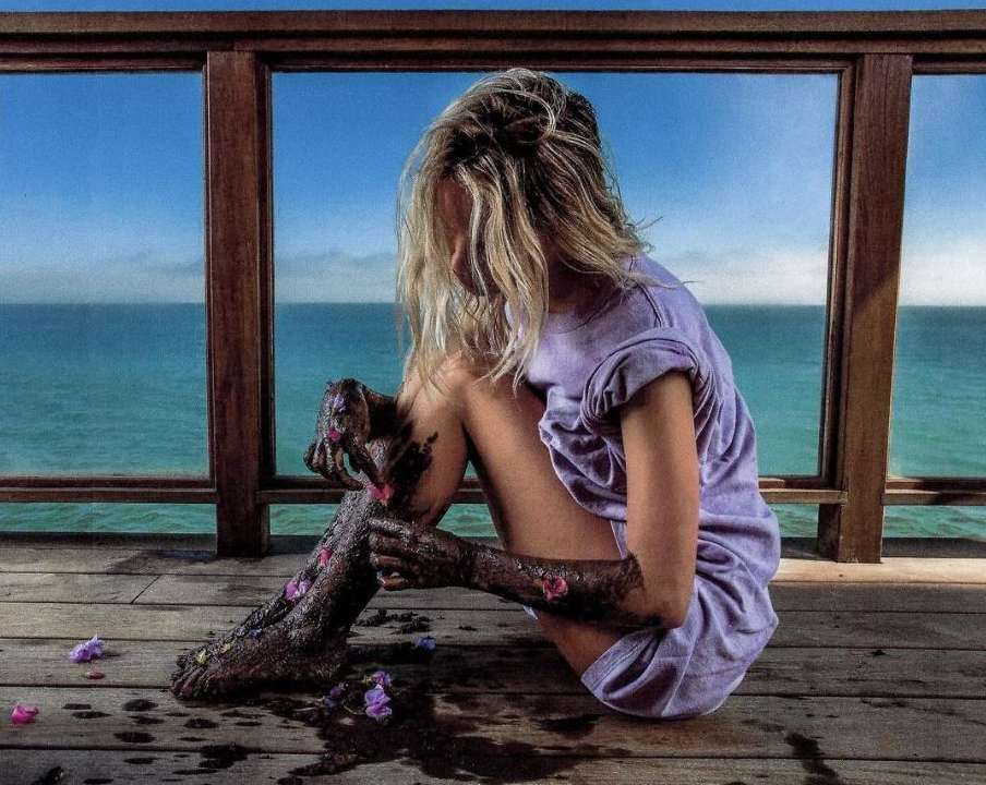 Woman seated on wooden pier covered in flowers, facing ocean horizon under blue sky. Woman in dress with blonde hair.