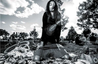 Black and white portrait of a woman kneeling on a patterned picnic blanket in a sunlit park by Brent Mckeever.