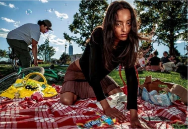 A woman kneeling on a red picnic blanket in a park with scattered items and people relaxing in the background, captured by Brent Mckeever