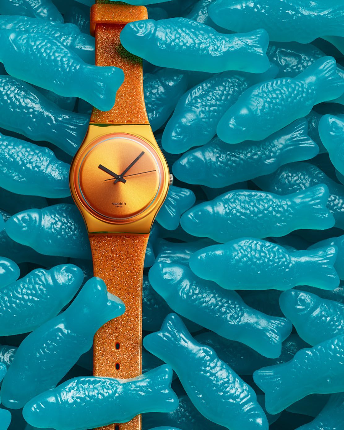 Close-up shot of an orange Swatch watch surrounded by many blue gummy fish candies, creating a playful and colourful visual.