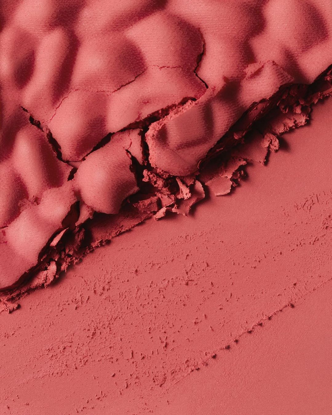 Macro photograph of a red powder blush with a cracked, uneven texture on one side and a smooth, pressed texture on the other.