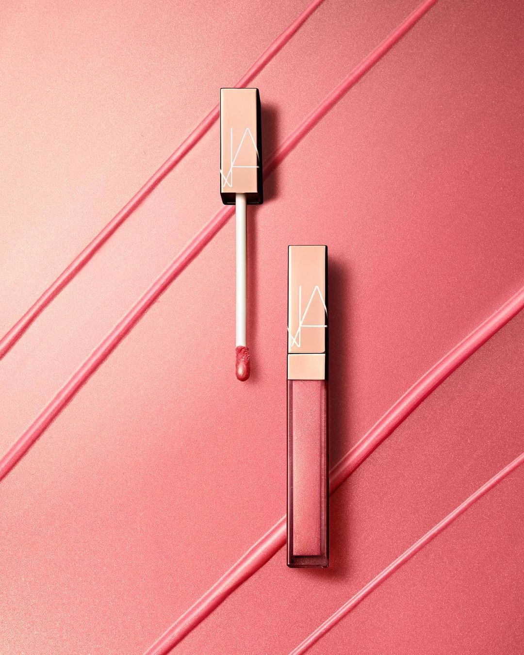 Two NARS lip glosses, one with applicator, on a pink surface with diagonal stripes of gloss. Beauty and cosmetics still life.