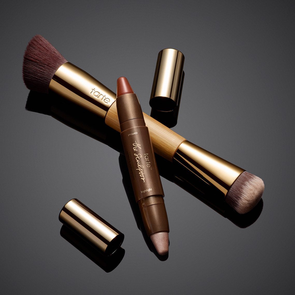 Tarte makeup products: a bronze-handled makeup brush and a dual-ended contour and highlighter stick, presented on a reflective surface.